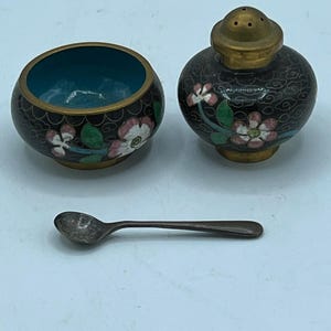Puede incluir: Un juego de sal y pimienta vintage con una cuchara pequeña. Los saleros y pimenteros son negros con diseños florales en rosa, blanco y verde. El salero tiene un interior turquesa y un borde dorado. La cuchara es plateada.
