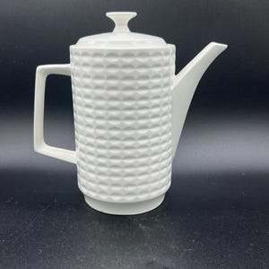 May include: A white ceramic coffee pot featuring a cylindrical body with a textured, repeating geometric pattern of small, raised semi-circles. The pot includes a matching lid with a small knob, a tall angular spout, and a square-shaped handle.