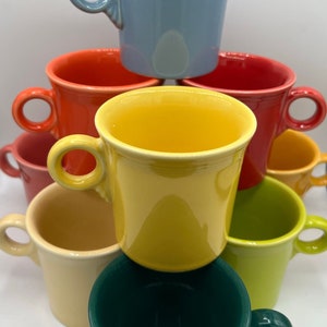 May include: A stack of colorful mugs in various shades of blue, orange, yellow, green, and cream. The mugs are all the same size and shape, with a simple, rounded design. They are made of ceramic and have a glossy finish.