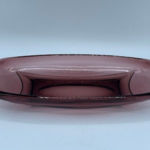 May include: A rectangular glass dish in a translucent plum purple color. The dish features a shallow basin with curved sides and rounded corners, showcasing a smooth, polished surface that reflects light across its transparent structure.