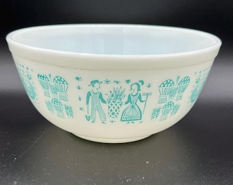 May include: Off-white mixing bowl with a turquoise design featuring a couple, baskets, and floral elements. The bowl has a light blue interior and a rounded shape. A vintage kitchenware item.