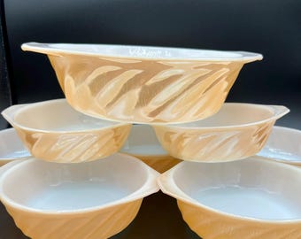 May include: A stack of vintage, peach-colored glass bowls with a white interior. The bowls have a distinctive swirl pattern on the exterior and small handles. The bowls are arranged on a dark surface.