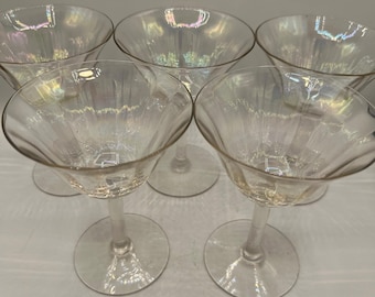May include: A set of five clear, iridescent champagne glasses. Each glass has a round base, a long stem, and a wide, fluted bowl. The glasses are arranged in a close-up shot, showcasing their elegant design and reflective qualities.