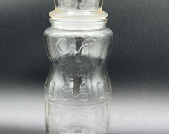 May include: Clear glass jar with a lid, featuring the words "MR PEANUT" on the lid. The jar has a unique shape with a wider base and a narrower middle section. The jar is empty and appears to be vintage.