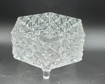 May include: A clear, faceted glass trinket box with a geometric pattern. The box is hexagonal with a lid and sits on a single, short foot. The glass reflects light, creating a sparkling effect.
