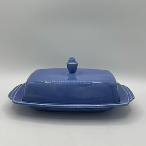 May include: A light blue ceramic butter dish with a lid. The dish has a rectangular shape with rounded edges and a small handle on the lid. The base has a scalloped design. The butter dish is a solid, pastel blue color.