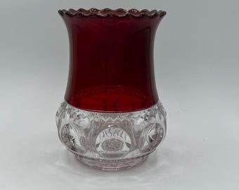 May include: A decorative glass vase with a deep red upper section and a clear, patterned lower section. The rim has a scalloped edge. The vase is designed with a bulbous base and a flared top, showcasing a vintage aesthetic.