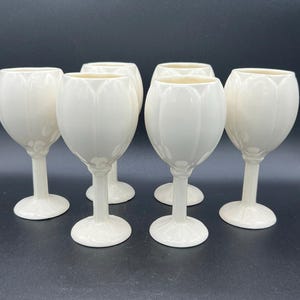 May include: A set of five off-white ceramic goblets, each featuring a smooth, rounded bowl with vertical petal-like indentations, a slender stem, and a circular base. The goblets have a glossy finish and are arranged in a group on a dark surface.