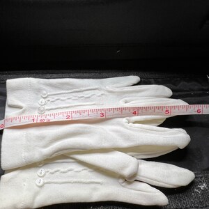 May include: A pair of white cotton gloves with delicate stitching and button closures. The gloves are laid out flat on a black surface with a measuring tape.