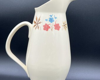 May include: Cream-colored ceramic pitcher with a curved handle and spout. The pitcher is decorated with a floral pattern in shades of pink, blue, and brown. A vintage-style water pitcher for serving beverages.