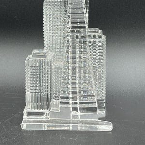 May include: A clear glass skyscraper sculpture with detailed architectural features. The building has multiple rectangular sections and a textured surface, creating a realistic representation of a city skyline. The sculpture is displayed against a dark background.
