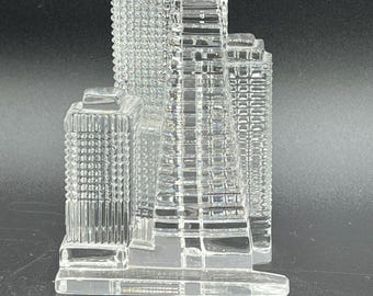 May include: A clear glass skyscraper sculpture with detailed architectural features. The building has multiple rectangular sections and a textured surface, creating a realistic representation of a city skyline. The sculpture is displayed against a dark background.