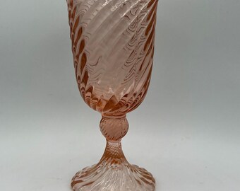 May include: A pink glass goblet with a spiral design. The goblet has a wide, flared rim and a detailed stem. The base is round and also features a spiral pattern. The glass has a translucent quality.