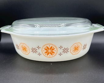 May include: A white, oval casserole dish with a clear glass lid. The dish features a decorative band of orange and brown geometric patterns. The lid has green accents on the handles. A vintage kitchenware item.