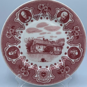 May include: A decorative plate with a red and white design. The plate features a central image of a building and portraits around the rim. Floral and scrollwork patterns adorn the border. The plate is displayed on a clear stand.
