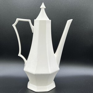 May include: A white ceramic coffee pot with a geometric design. The pot has a tall, faceted body, a long spout, and a handle with a unique, angular shape. The lid is conical, and the base is octagonal.