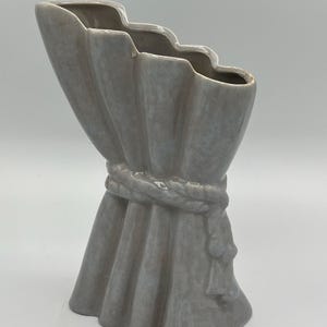 May include: A gray ceramic vase with a unique draped design. The vase has a wavy top edge and is cinched in the middle with a rope-like detail. The bottom flares out, creating a sculptural and decorative piece.