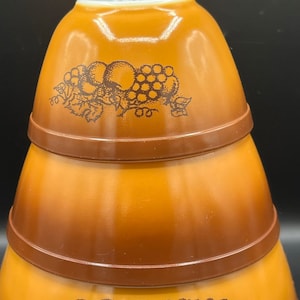 May include: Set of three brown ceramic bowls with a grape and apple design. The bowls are stacked on top of each other.