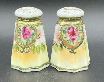 May include: A pair of antique salt and pepper shakers. Each shaker features a white, gold, and yellow design with a floral motif. The shakers have a shield-shaped design with pink roses and green leaves.
