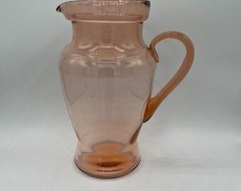 May include: A vintage pink glass pitcher with a handle and a spout. The pitcher has a rounded body and a flared base. The handle is curved and attached to the side of the pitcher. The pitcher is made of clear glass and has a smooth surface.