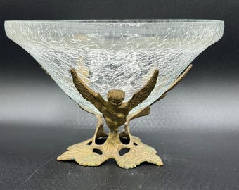 May include: A clear glass bowl with a textured surface, supported by a bronze-colored metal base. The base features a stylized bird with outstretched wings and a leaf-like design. The bowl is likely for decorative purposes.