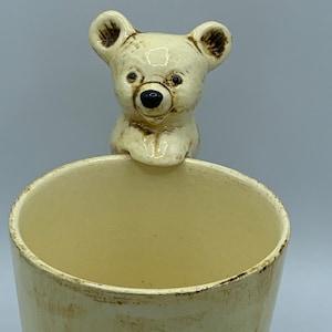 May include: A cream-colored ceramic mug with a bear-shaped handle. The bear is white with brown eyes and a black nose.