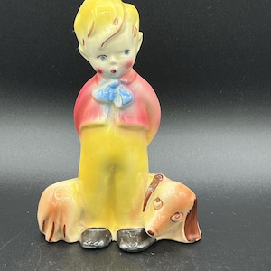 May include: A ceramic figurine of a young person with blonde hair, wearing a red jacket, blue necktie, and yellow trousers, standing beside a brown and tan dachshund dog. The figure has painted facial features and black shoes.