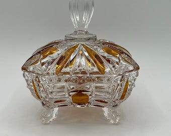 May include: A clear, cut-glass candy dish with a lid and a decorative handle. The dish features amber-colored accents and sits on three small feet. The lid has a clear, elongated handle. The dish is designed with intricate geometric patterns.