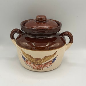 May include: A brown ceramic crock with a lid, featuring a painted image of an eagle with American flags. The text "The Spirit of" and "Freedom" is visible on the crock.