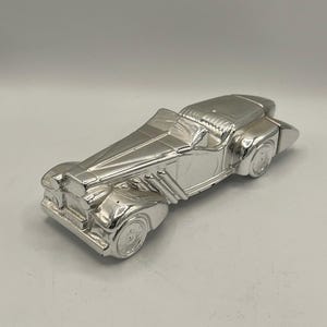 May include: A silver-colored, vintage-style car figurine. The detailed model features a long hood, open cockpit, and prominent fenders. The car has a classic design, with visible wheels and exhaust pipes, and is made of a shiny, reflective material.