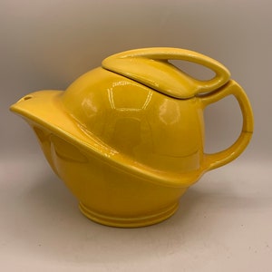 May include: A yellow ceramic teapot with a unique, modern design. The teapot has a rounded body and a spout that curves upwards. The lid has a handle that is shaped like a loop.
