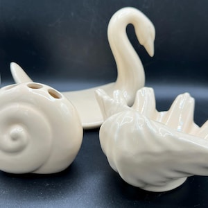 May include: Three off-white ceramic decorative pieces: a swan, a shell-shaped item, and a bird-shaped item. The swan has a long neck and is in a resting position. The shell-shaped item has three holes. The bird-shaped item has a curved shape.