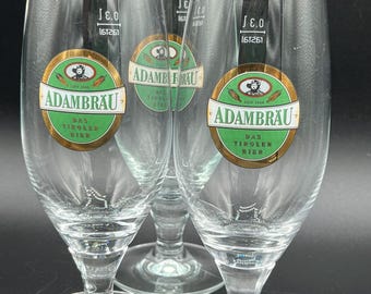 May include: Clear stemmed beer glasses with the "ADAMBRAU" logo in green and gold. The glasses have a wide bowl and a slender stem. The text "DAS TIROLER BIER" is also visible on the label. The glasses are arranged in a group.