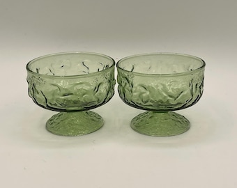 May include: Two green glass dessert bowls with textured, organic patterns. Each bowl has a rounded base and a pedestal foot. The bowls are made of a translucent green glass, and the texture gives them a unique, vintage look.