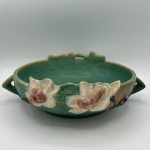 May include: A round ceramic bowl featuring a teal and green glazed finish with two side handles. The exterior is decorated with raised, three-dimensional white and pink floral motifs. The rim has a scalloped edge with small cut-out details.
