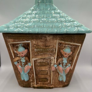 May include: A ceramic cookie jar shaped like a house with a blue roof and two elf figures on the sides. The house has a brown door with a sign that says "Welcome".