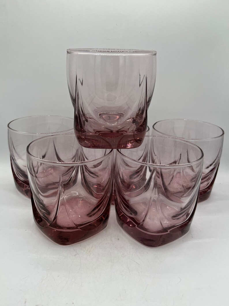 May include: Set of six clear glass tumblers with a pink hue and a square shape. The glasses have a decorative design with a wavy pattern.