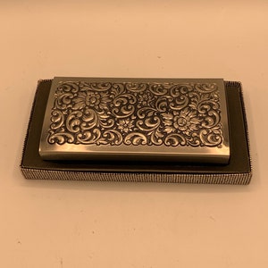 May include: A silver metal box with an ornate floral design. The box is closed and sitting on a black surface.