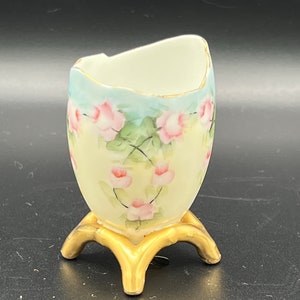 Puede incluir: Un pequeño jarrón de porcelana en forma de huevo con un diseño floral en tonos rosa, verde y azul. El jarrón tiene una base pintada en oro con tres patas.