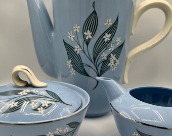 Könnte beinhalten: Ein blaues und weißes Blumen-Tee-Set mit Teekanne, Milchkännchen und Zuckerdose. Das Set zeigt ein zartes Blumenmuster mit weißen Blüten und grünen Blättern auf hellblauem Hintergrund.