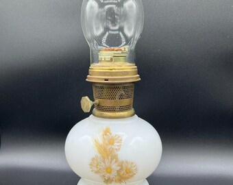 May include: A vintage oil lamp with a clear glass chimney and a white base. The base features a floral design in yellow and white. The lamp has a brass-colored metal top and a knob for adjusting the wick. The lamp is designed for kerosene or oil.