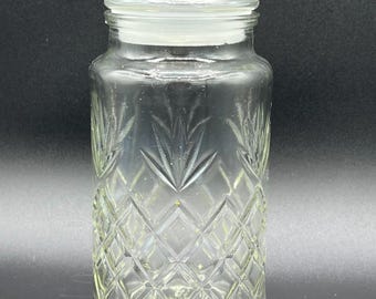 May include: A clear glass jar with a lid. The jar has a diamond pattern on the lower half and a leaf design on the upper half. The lid is flat and clear, and the jar is cylindrical. The jar is empty and sits on a black surface.