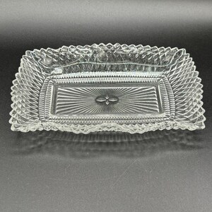 May include: A clear, rectangular glass dish with a decorative, diamond-patterned border. The center features a sunburst design with a small, floral-like detail. The dish is likely for serving or display.
