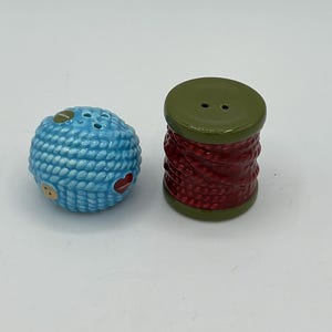 May include: A ceramic salt and pepper shaker set featuring a round, light blue ball textured like a ball of yarn with small button accents, and a cylindrical, dark red spool of thread with olive green ends, both with small holes on top.