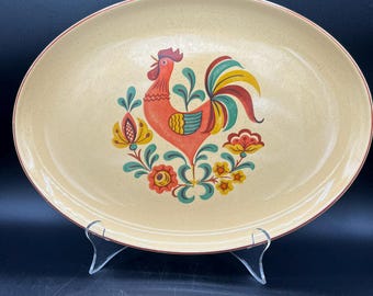 May include: Oval serving platter with a rooster design. The beige platter features a red, orange, yellow, green, and blue rooster illustration in the center. The rim is a darker red. The platter is displayed on a clear stand.
