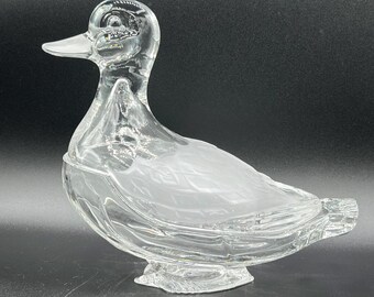 May include: A clear glass duck figurine with a detailed head, beak, and body. The duck has a textured, frosted design on its back and is mounted on a decorative base. The figurine is a decorative item.