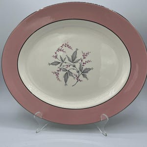 May include: An oval ceramic serving platter featuring a wide dusty pink border and a silver-toned rim. The center of the platter is cream-colored and decorated with a central botanical illustration of grey leaves and small pink floral accents.