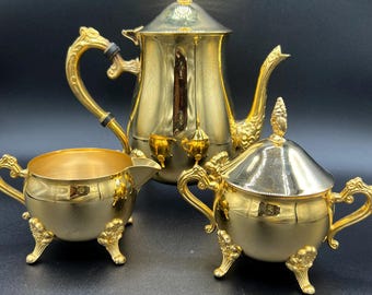 May include: A gold-colored tea set with ornate detailing. The set includes a teapot with a lid, a creamer, and a sugar bowl with a lid. Each piece has decorative handles and rests on small, detailed feet. The set is displayed against a dark background.