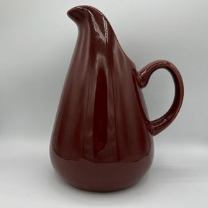May include: A maroon-colored ceramic pitcher with a curved spout and a rounded body. The pitcher has a handle and a glossy finish, suitable for serving beverages. The design is simple and elegant, suitable for any kitchen.
