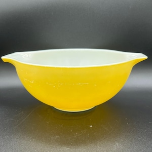 May include: A vintage yellow mixing bowl with a white interior. The bowl has a rounded shape with two small handles on either side. The bowl is made of glass and has a glossy finish.